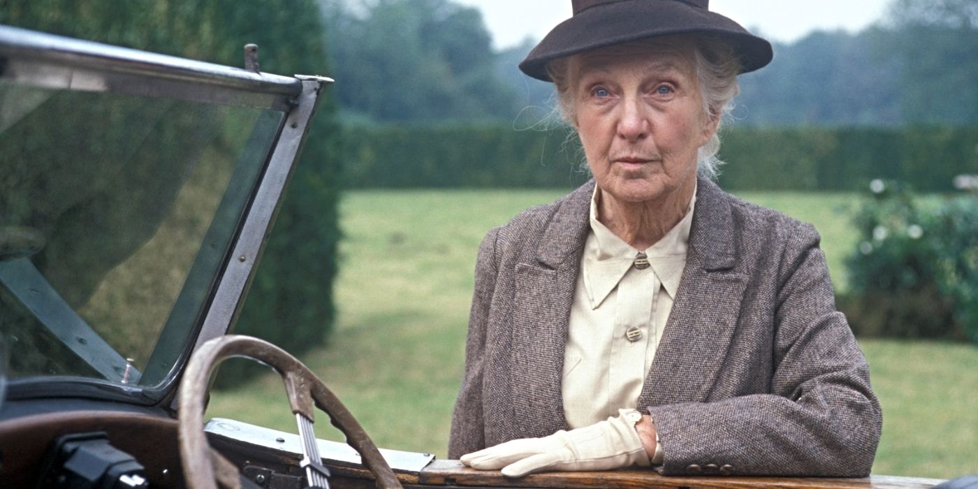 Joan Hickson as Miss Marple, wearing a hat and gloves and leaning against a car in Miss Marple