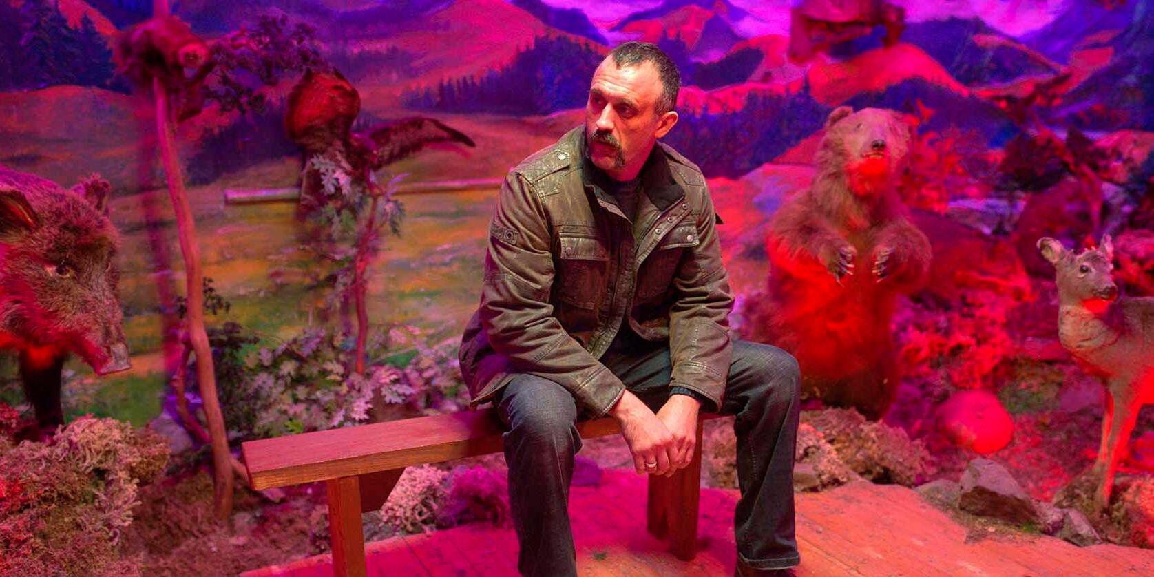 A still from the film Pamfir featuring a man sitting on a bench surrounded by diorama woodland creatures