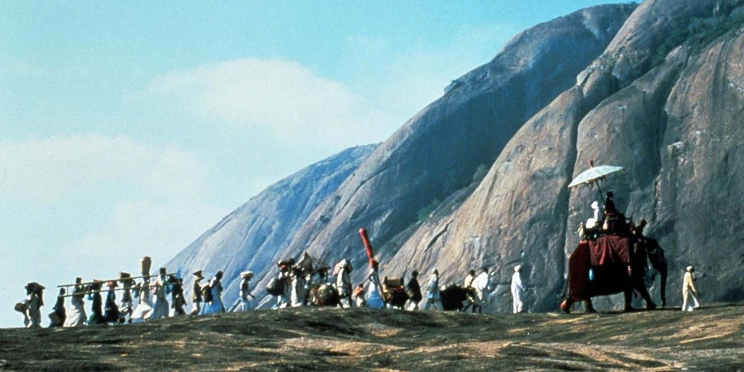 people walking and two people riding an elephant in front of a mountain