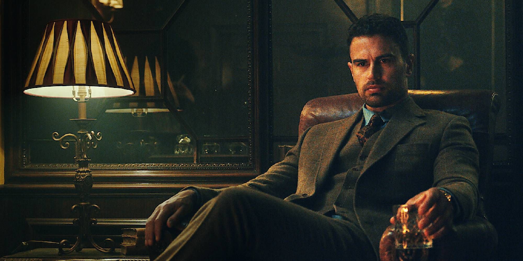 Theo James in The Gentlemen sitting in a chair and holding a glass of whiskey.