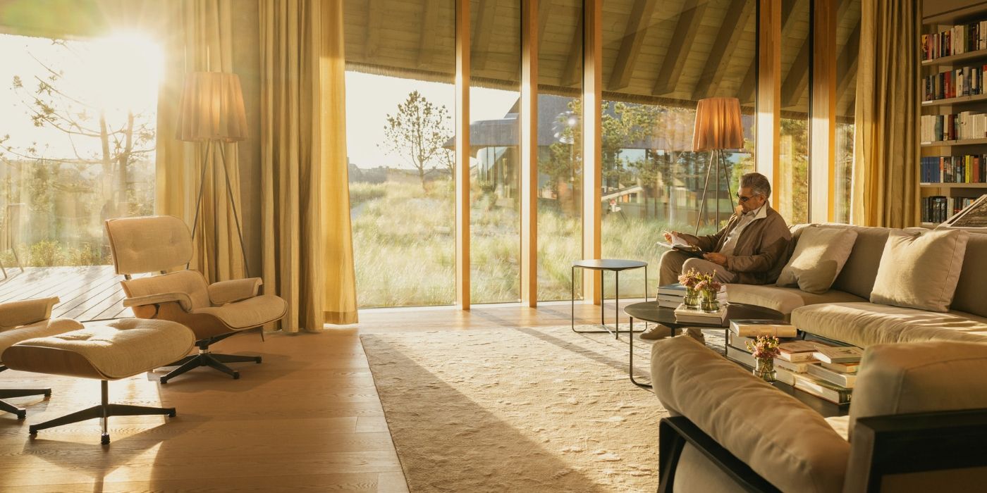 Eugene Levy se sienta en un sofá en una habitación soleada de un resort alemán en la segunda temporada de The Reluctant Traveler