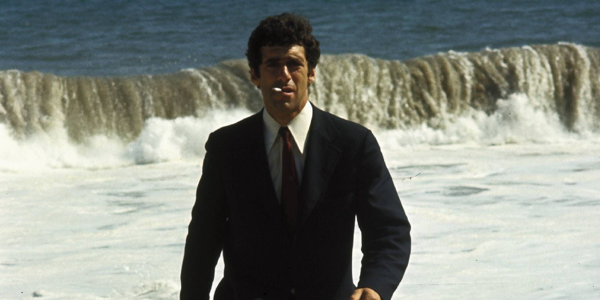 Elliott Gould in a suit smoking a cigarette at the beach while waves splash behind him