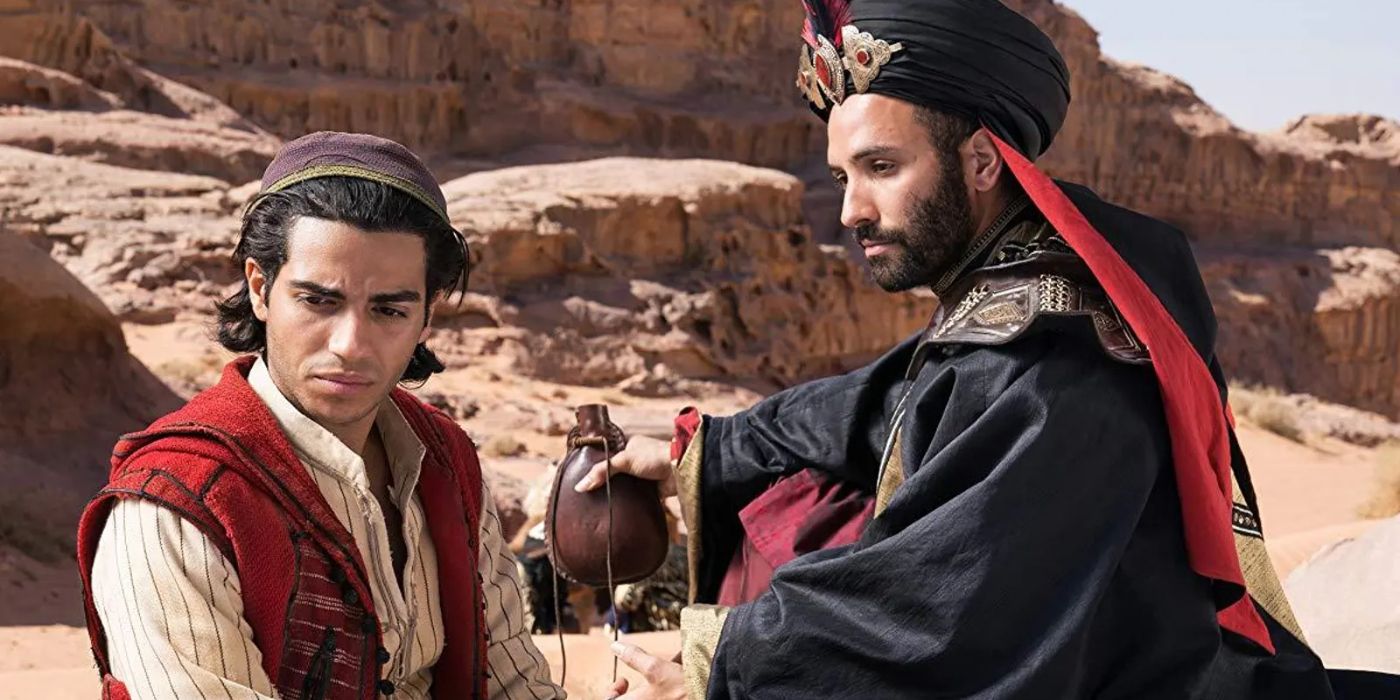 Aladdin (Mena Massoud) sits in the desert with Jafar (Marwan Kenzari) who holds up a water bottle in 'Aladdin' (2019).