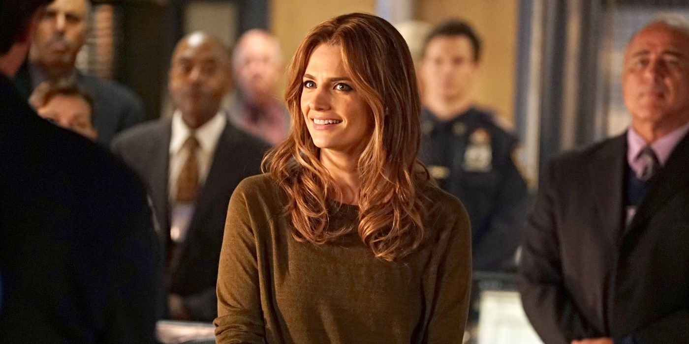 Stana Katic as Kate Beckett, wearing a brown sweater and smiling while a group of people stand behind her on Castle