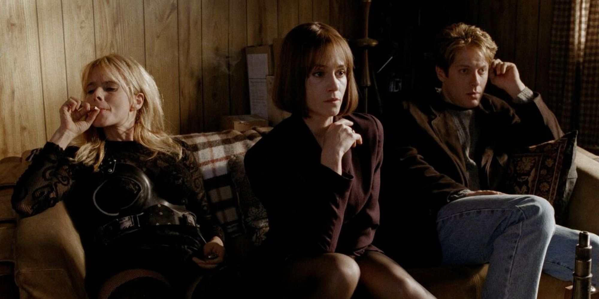 Three people sit on a couch, two look blankly ahead while one smokes a cigarette in Crash, 1996.