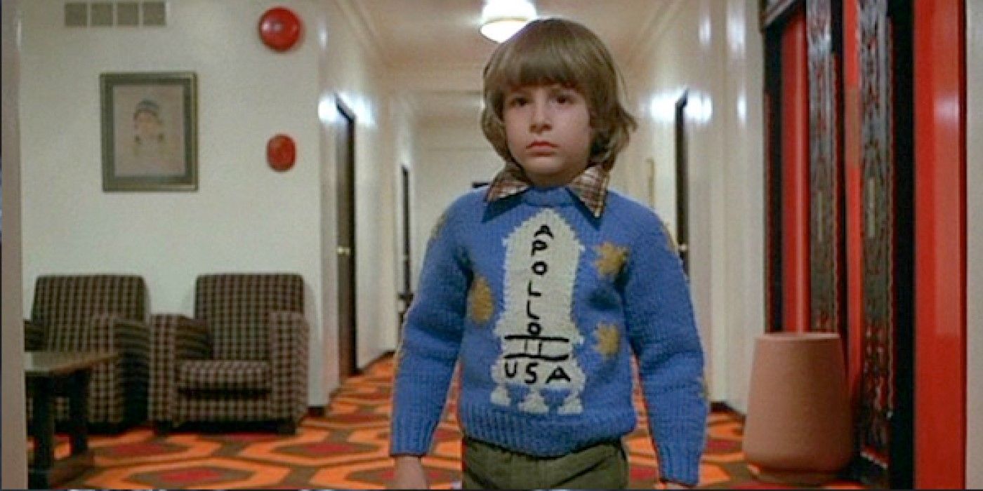 Young Danny Torrance (Danny Lloyd) stands facing forward in a hallway of the Overlook Hotel, wearing a blue Apollo 11 sweater in 'The Shining'