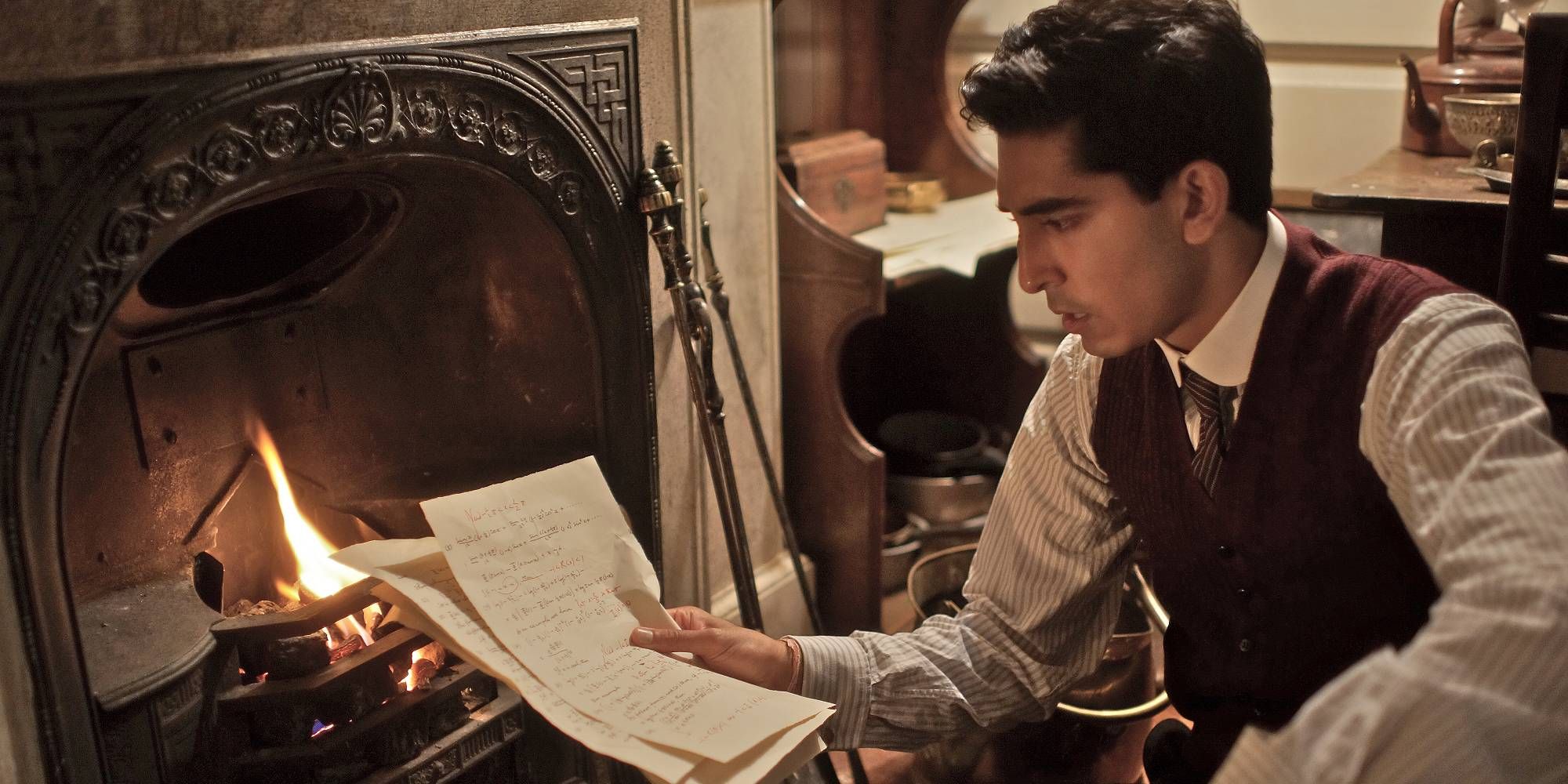 Dev Patel de El hombre que conoce el infinito está leyendo un periódico junto a la chimenea.