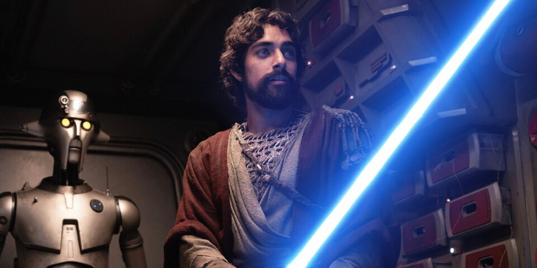 A bearded Ezra Bridger holds a blue lightsaber with a robot behind him in 'Ahsoka' on Disney+