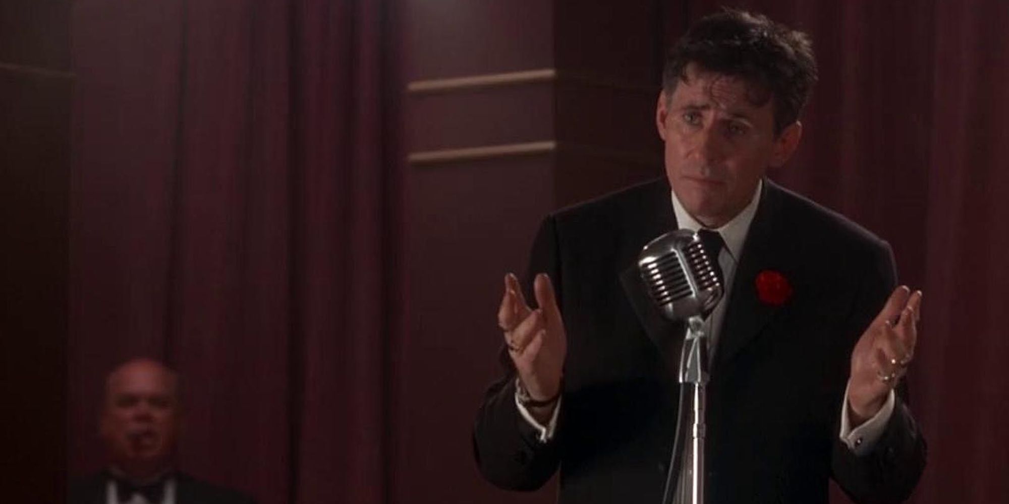 A man in a suit stands with his hands out behind a microphone stand in Mad Dog Time