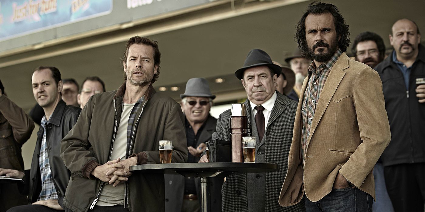 Guy Pearce and Aaron Pedersen standing around high top tables and looking out in Jack Irish