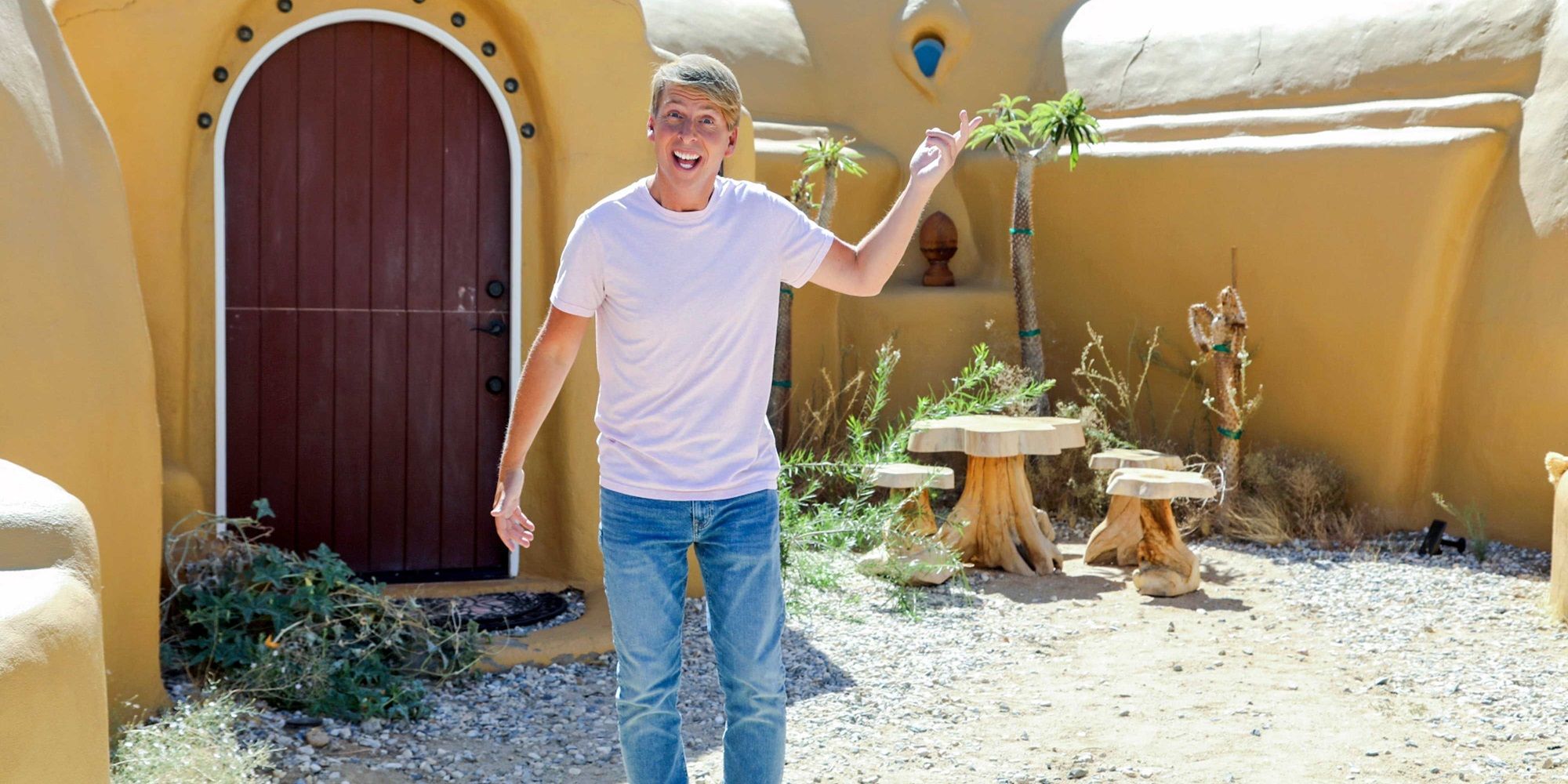 Jack McBrayer, wearing a white T-shirt and blue jeans while pointing at a yellow house, serving as the host of Zillow Gone Wild at HGTV