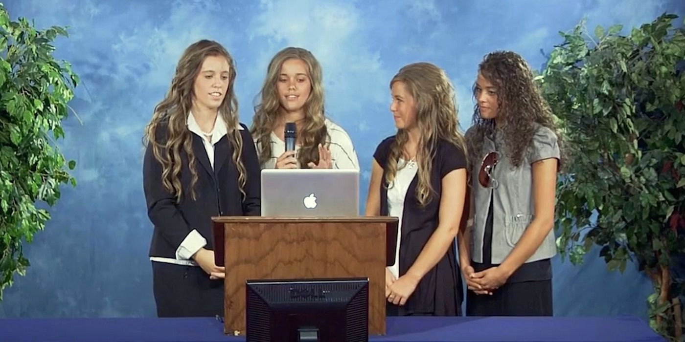 Jill Duggar stands at a podium reading from a laptop with three sisters standing with her