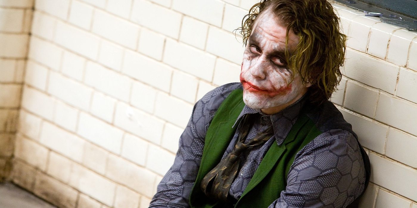 Joker (Heath Ledger) sits on the floor of an interrogation room with his back against the wall in The Dark Knight