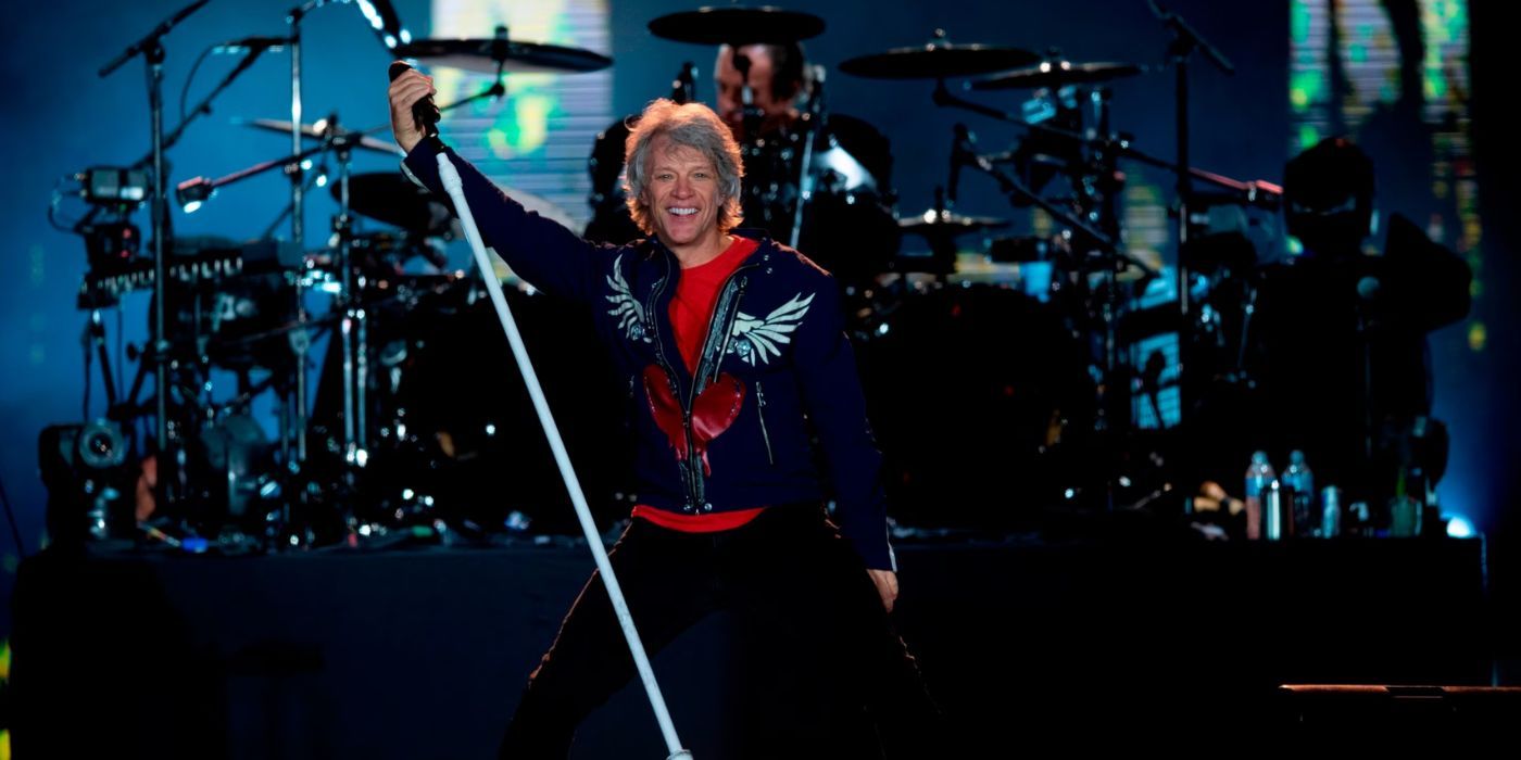 Jon Bon Jovi is holding a microphone stand in his right hand on a dark stage in 'Thank You, Goodnight: The Bon Jovi Story'