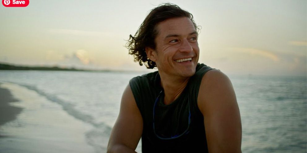 Orlando Bloom looking off to the side smiling on a beach with the ocean behind him