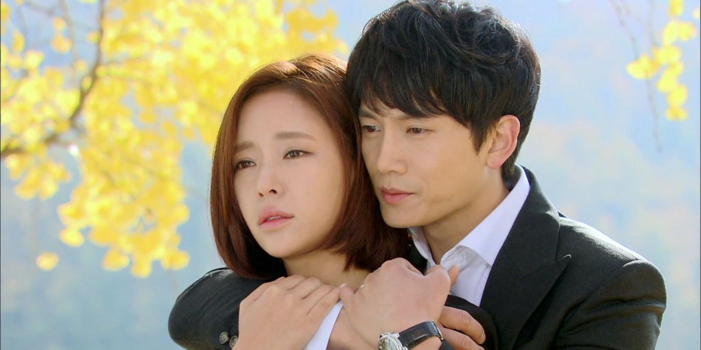 A man stands behind a woman and embraces her as they look to the distance with yellow trees in Secret Love.