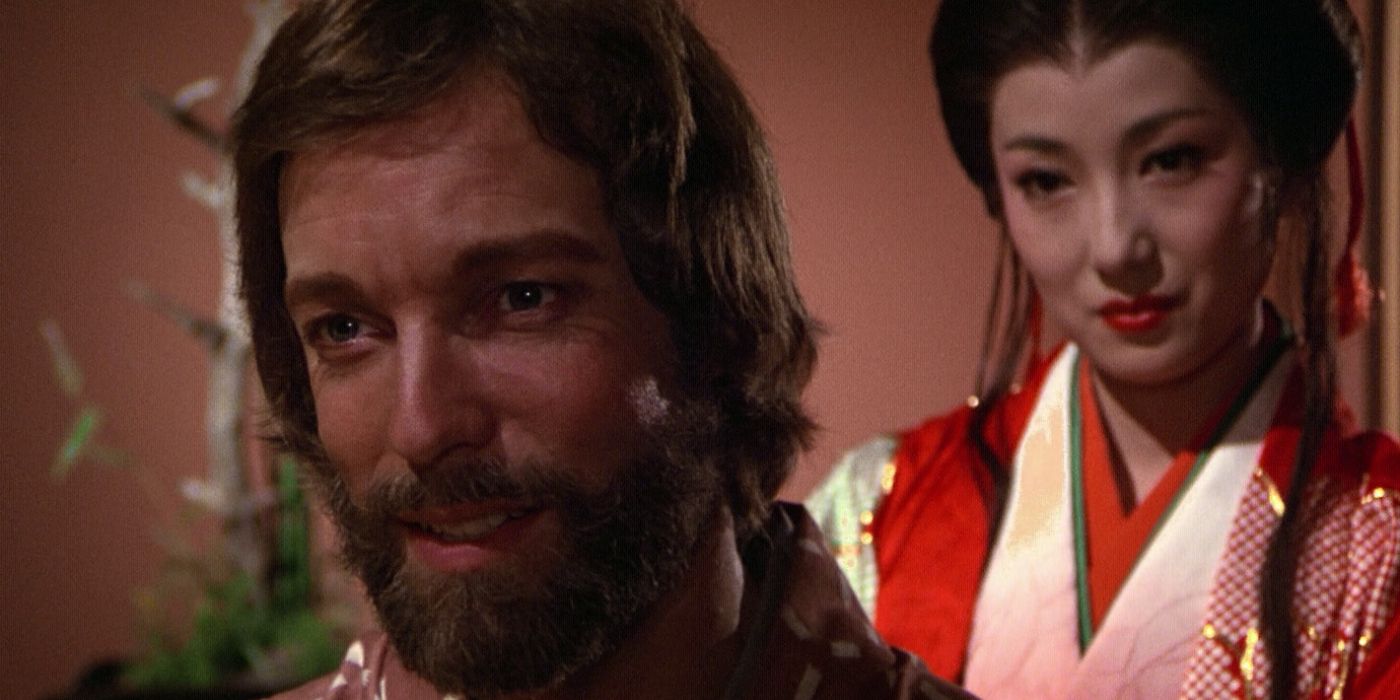 John Blackthorne (Richard Chamberlain) smiling and looking slightly to the left while sitting in front of a pink wall with a woman behind his right shoulder in the 1980 Shogun