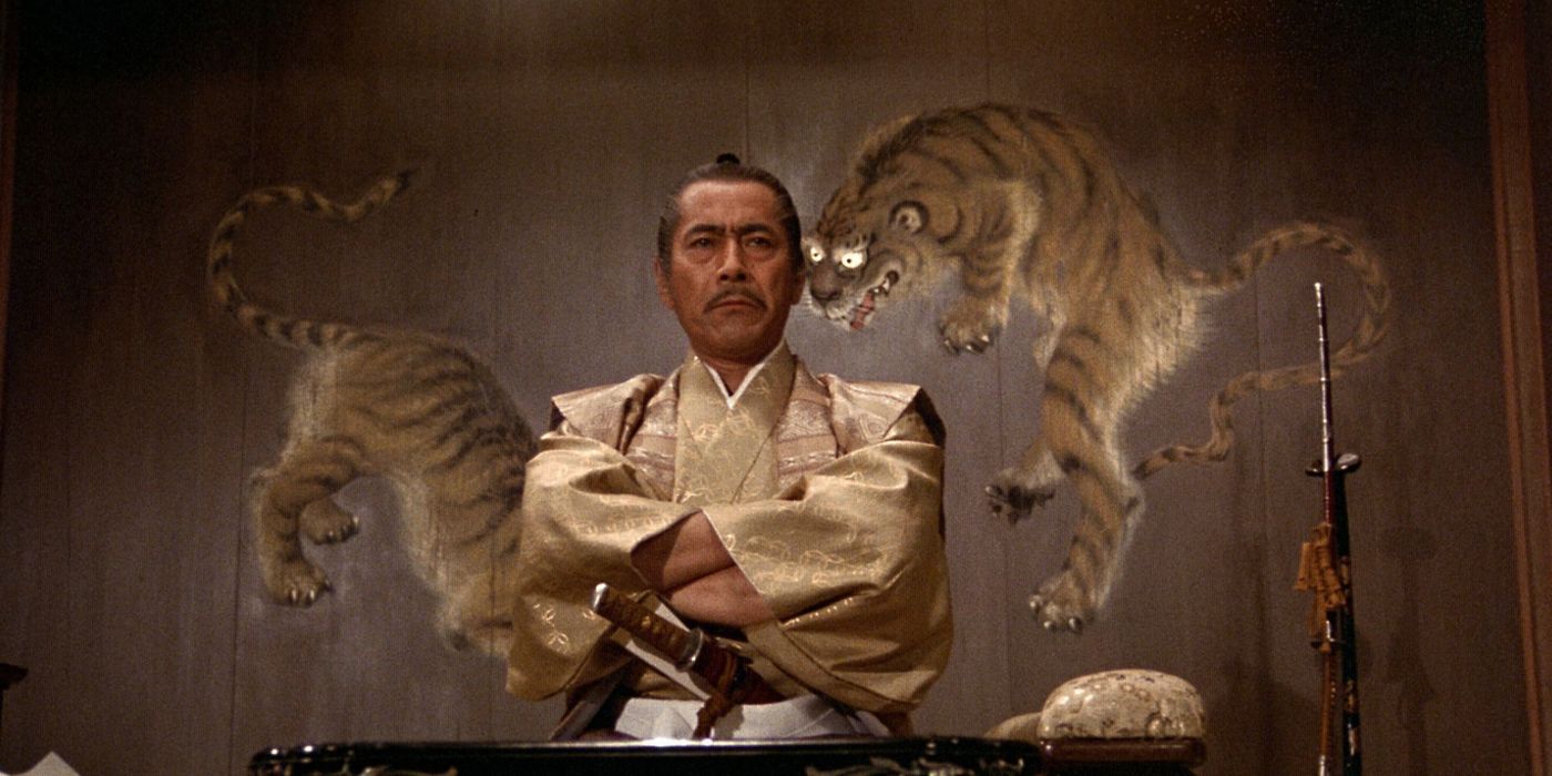 Toranaga (Toshiro Mifune) sitting down with crossed arms and looking sternly ahead with a brown-ish wall decorated with tiger art behind him in the 1980 Shogun