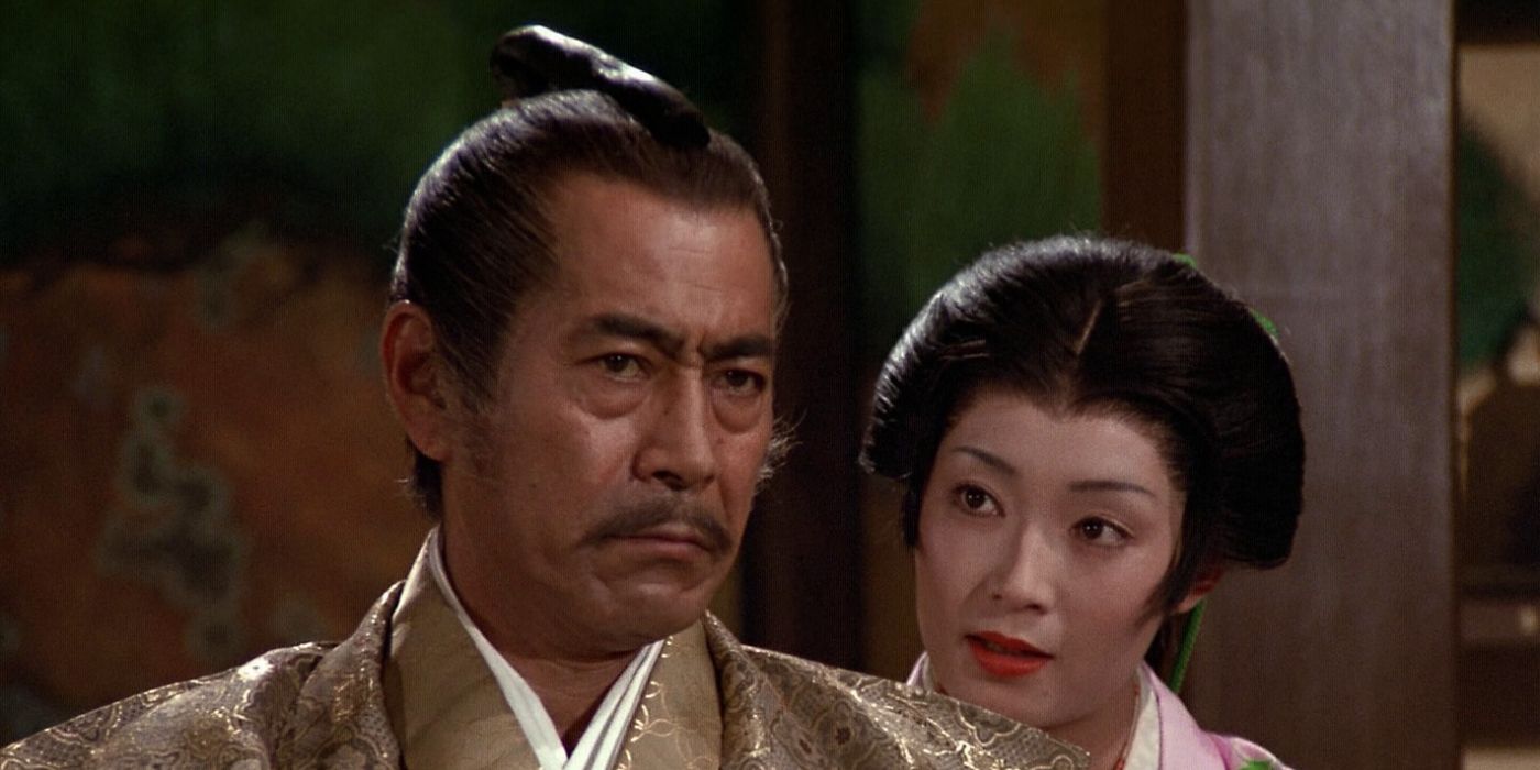 Toranaga (Toshiro Mifune) staring sternly ahead and slightly to the right with Mariko (Yoko Shimada) behind his right shoulder translating quietly in his ear in the 1980 Shogun