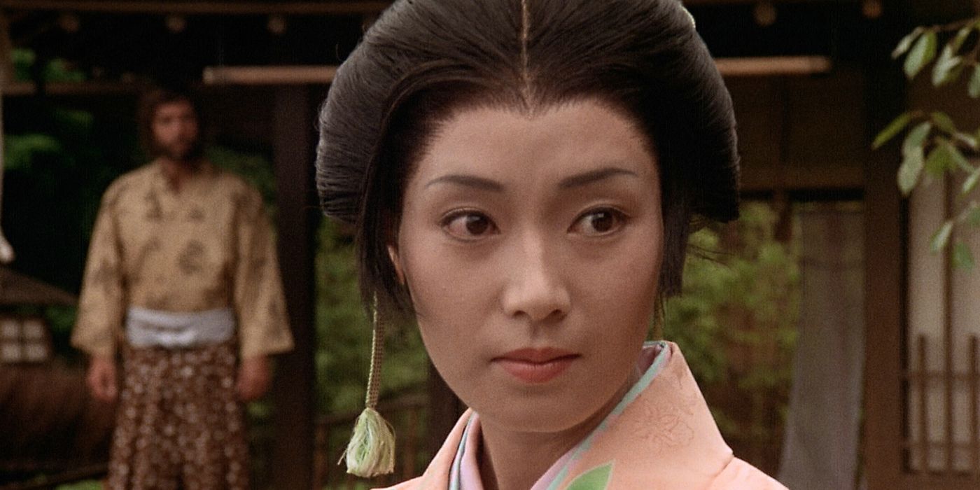 A close-up of Mariko (Yoko Shimada) looking slightly to the right with Blackthorne (Richard Chamberlain) standing behind her in the distance and out of focus in the 1980 Shogun