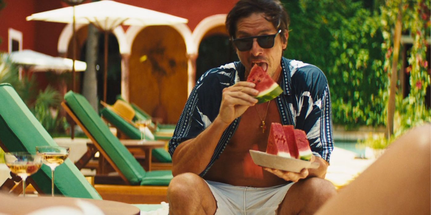 Simon Rex sitting on a pool chair eating watermelon slices with his shirt unbuttoned in Blink Twice.