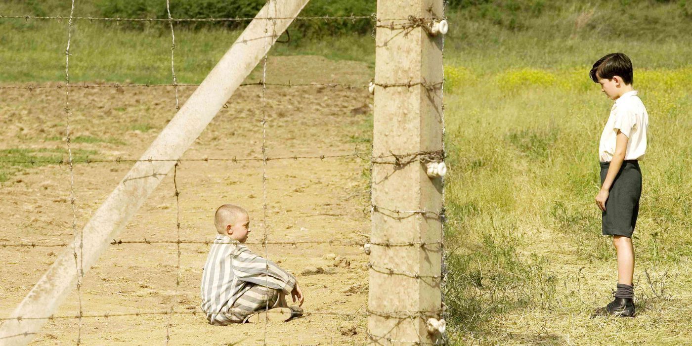 Shmuel (left) sits on the ground and talks to Bruno (right), who stands on the other side of a barbed wire fence