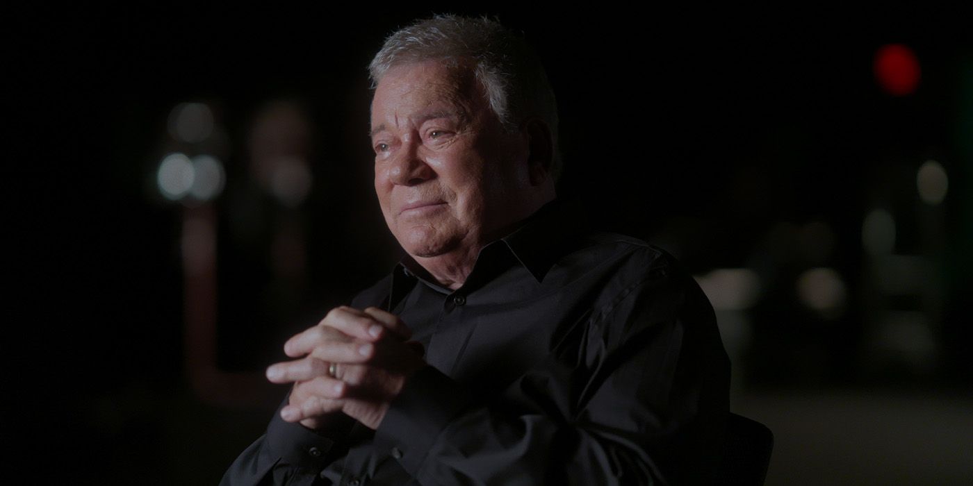 William Shatner smiles while looking off-camera with his hands clasped in front of him 