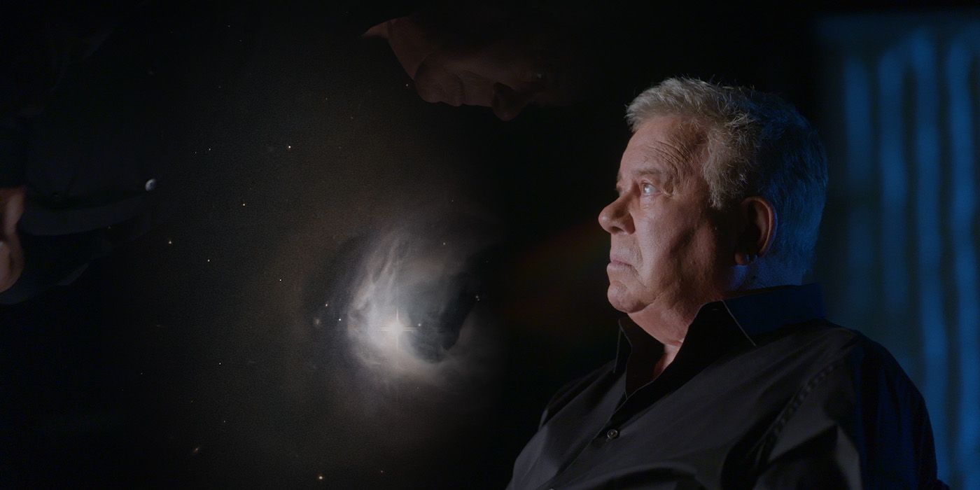 William Shatner sits looking up pensively towards a projection of space