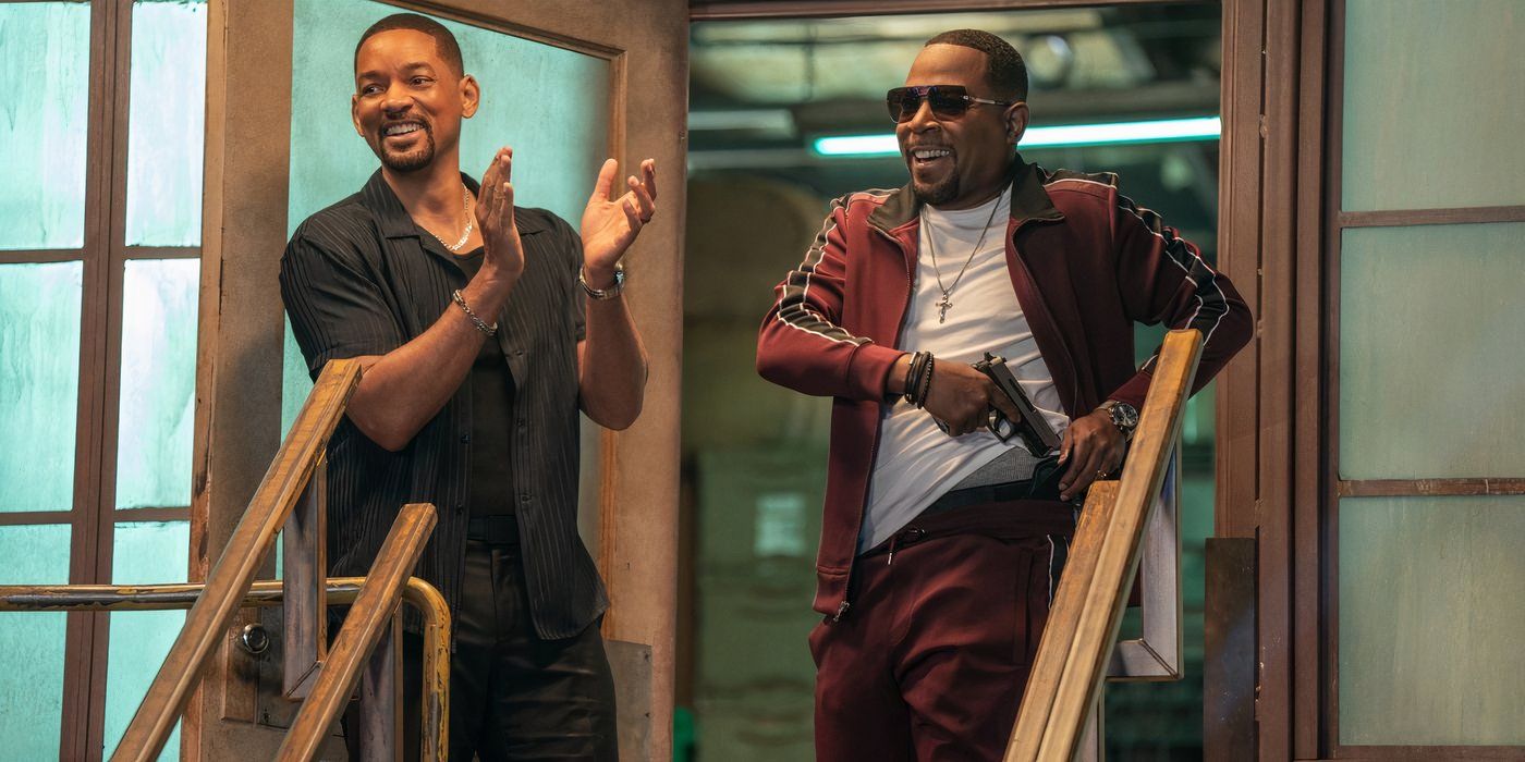 Will Smith smiling and clapping in front of a doorway next to Martin Lawrence wearing sunglasses and holstering his pistol.