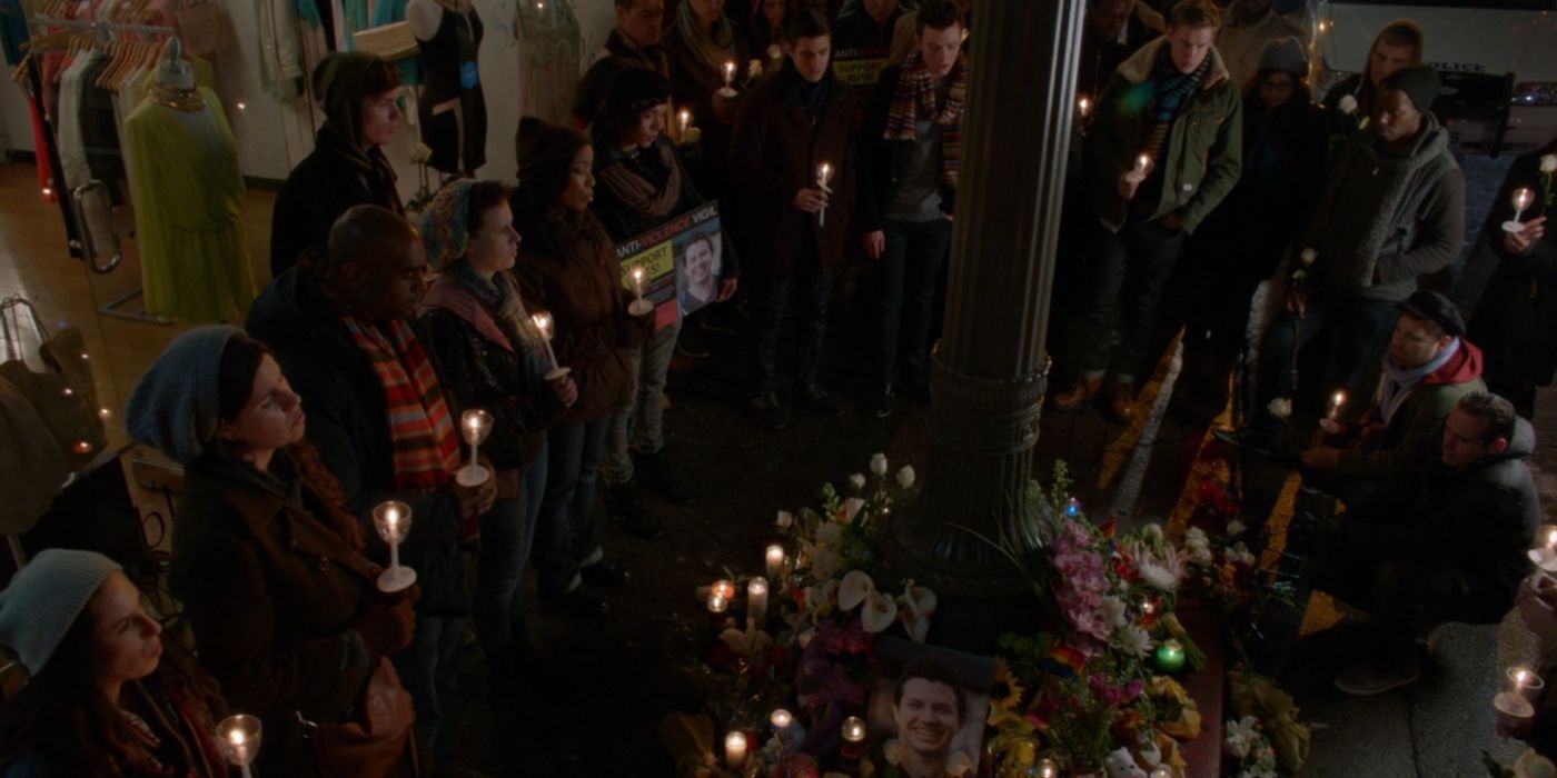 A group of people, including Rachel, Kurt, Blaine, and Sam gather around a memorial for someone who was killed in an attack. They are all holding candles. A picture of a brown-haired man smiling is surrounded by flowers, stuffed animals, and candles.