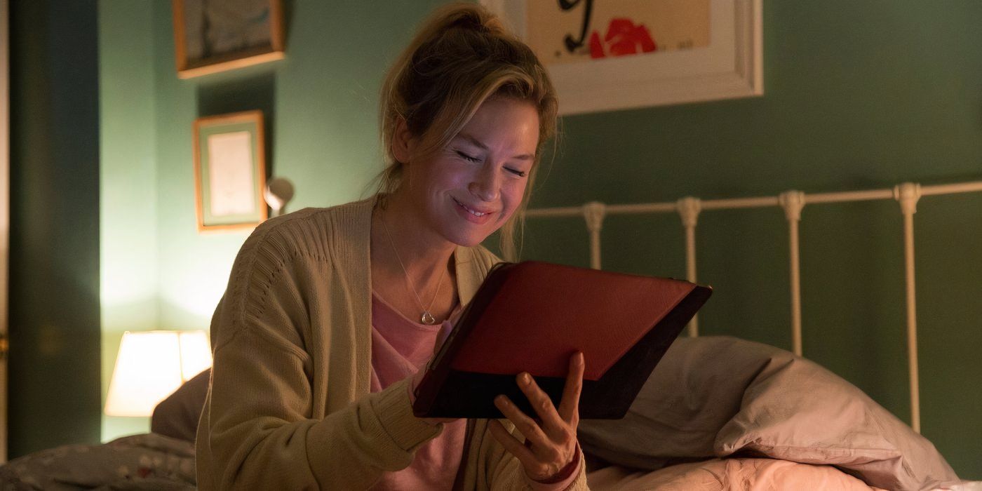 Renée Zellweger as Bridget Jones holding an iPad and smiling while sitting on a bed.