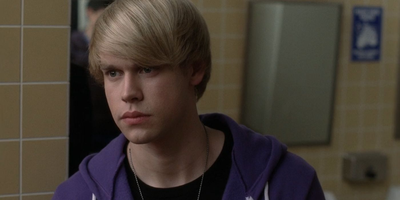 Sam, wearing a purple hoodie, black t-shirt, a silver chain, and jeans, intensely looks at Finn while in the bathroom at McKinley High. Sam's blonde hair is stylized similar to Justin Bieber's hair from early on in his career.