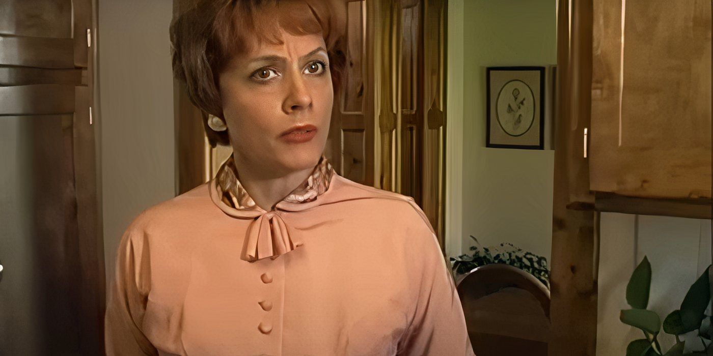 A woman in a pink blouse looks annoyed at the camera inside of her house
