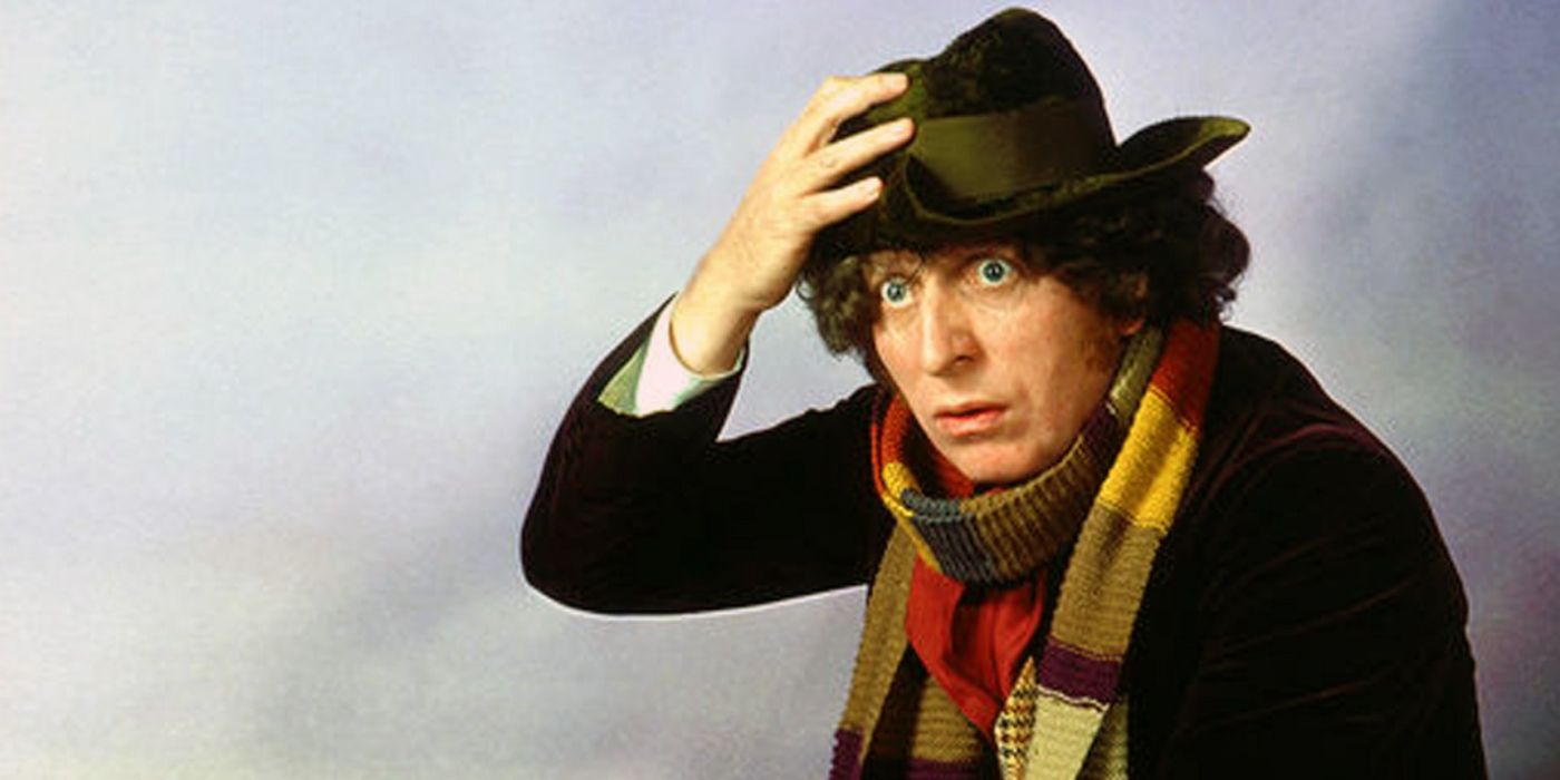 The Fourth Doctor (Tom Baker) standing against a white-gray backround clutching his hat to his head, wearing his scarf and coat, and looking offscreen to the left with a mildly perplexed expression, in Doctor Who
