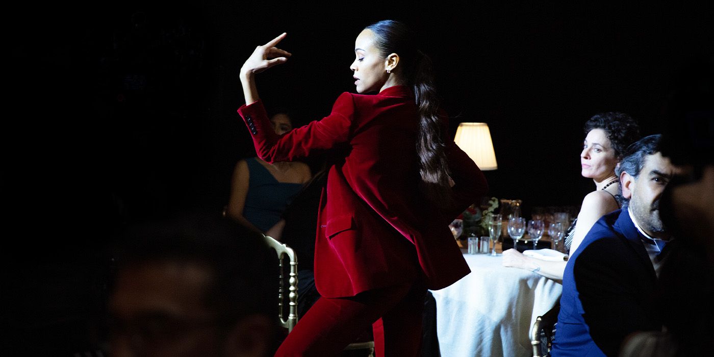 Zoë Saldaña dances in a red suit in a darkened room around tables of people