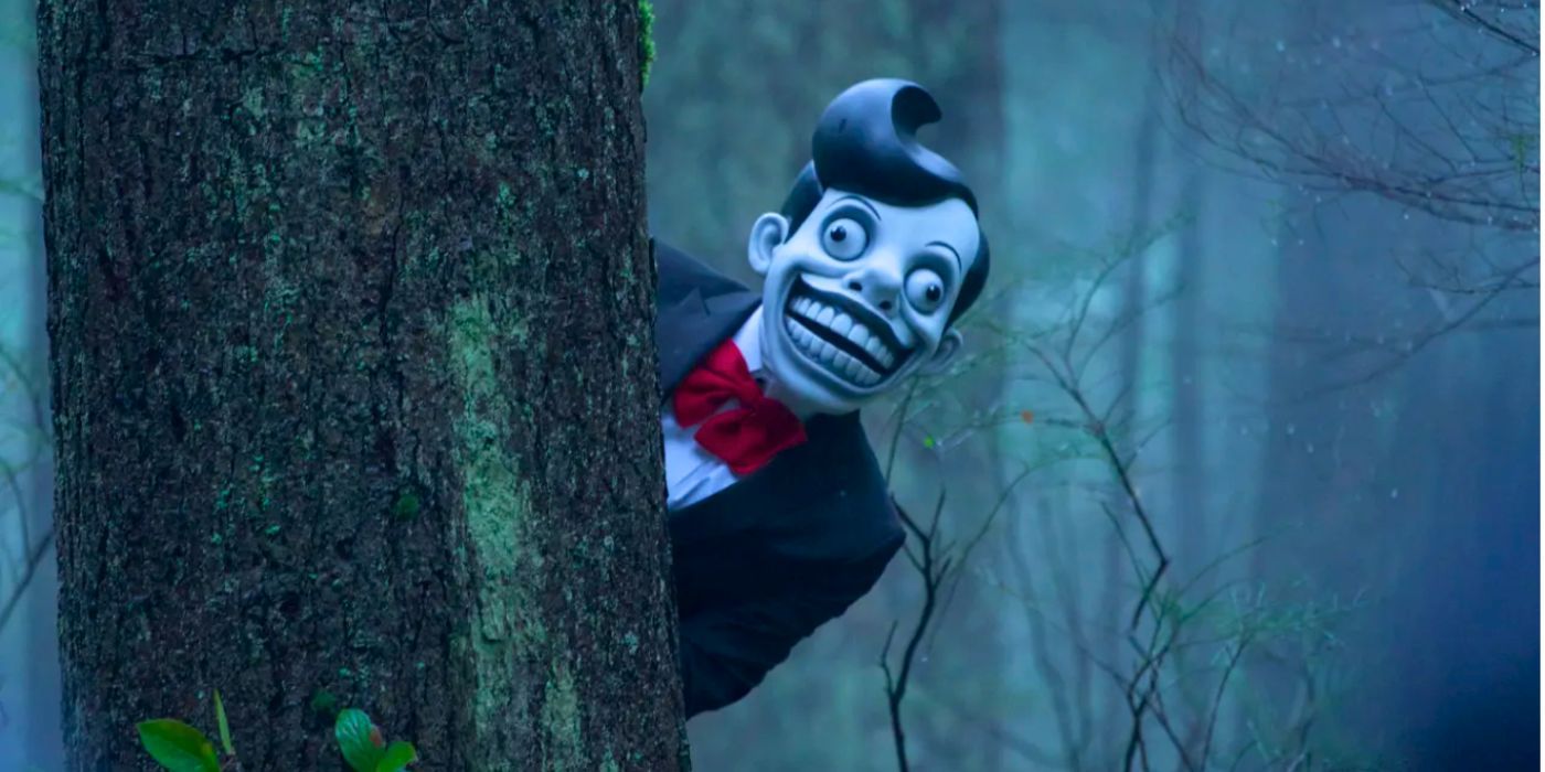 Creepy man wearing a toothy mask of fictional TV star Mr Chuckle Teeth hides behind a tree in the episode Familiar from The X-Files