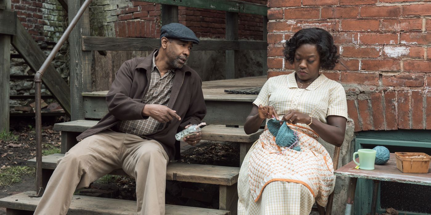 Denzel Washington holding a liquor bottle and sitting on some wooden steps next to Viola Davis who is knitting a wool cap.