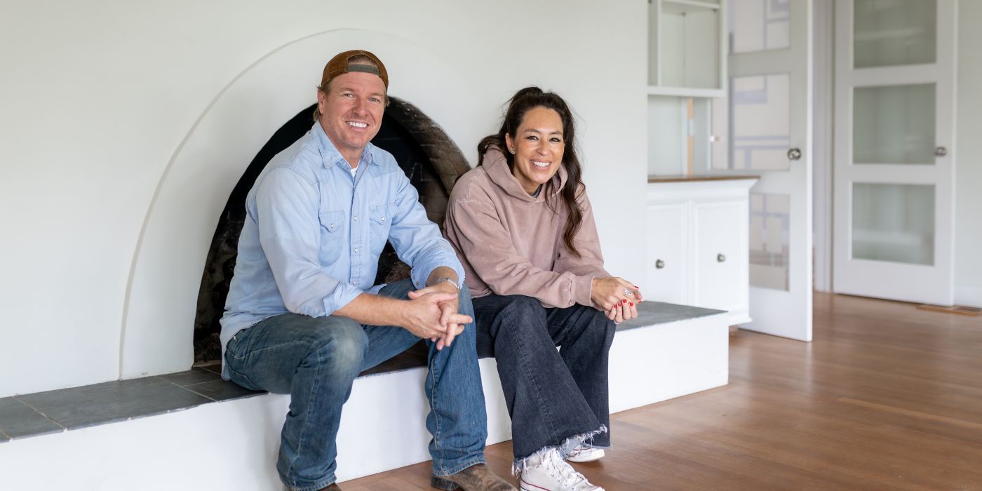 Chip and Joanna Gaines sitting in a white room on a stoop in front of a fireplace.