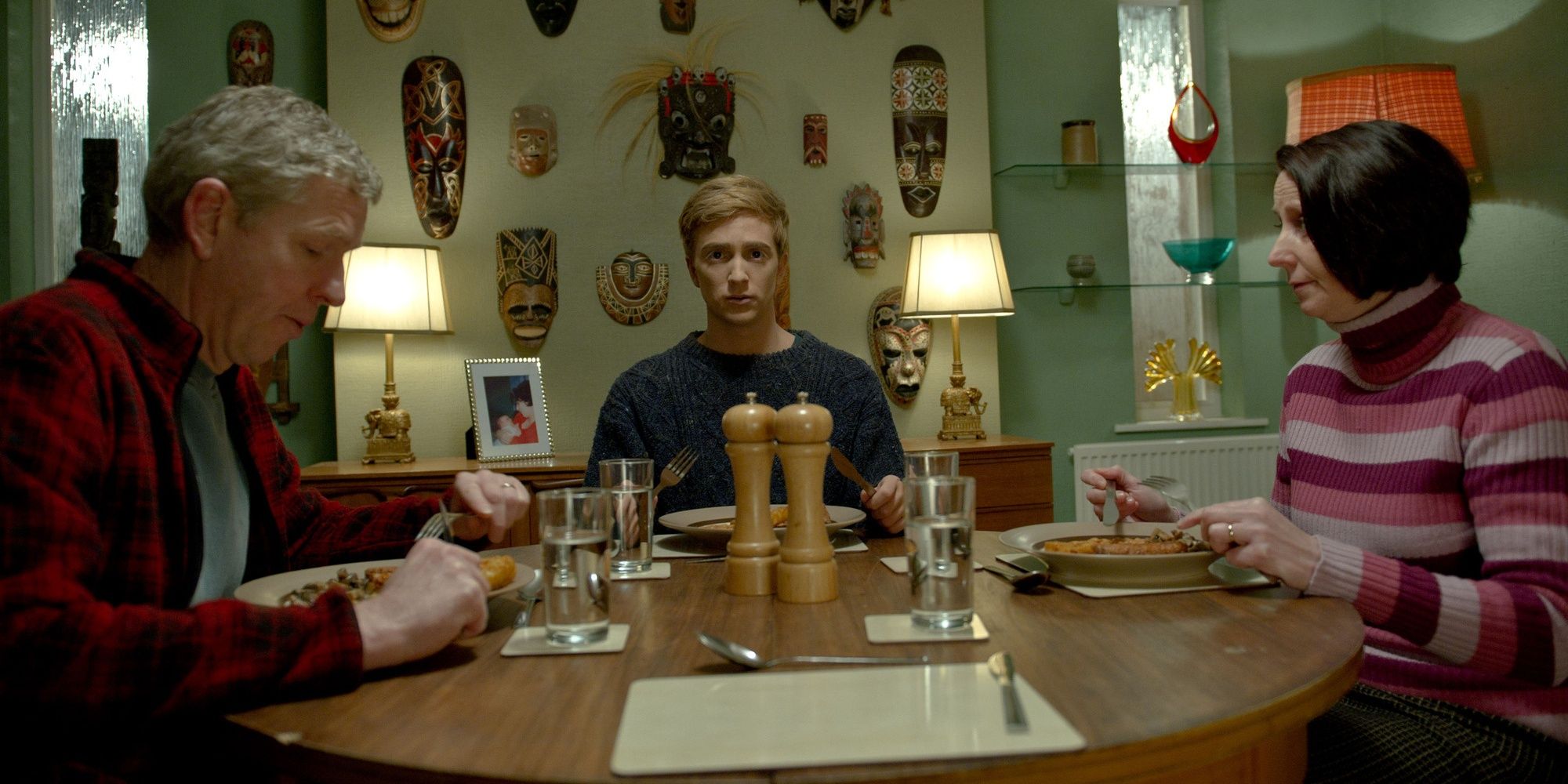 Steve Cooper, Luke Newberry and Marie Critchley sit at a dinner table surrounded by masks as Steven, Kieren and Sue Walker in In the Flesh