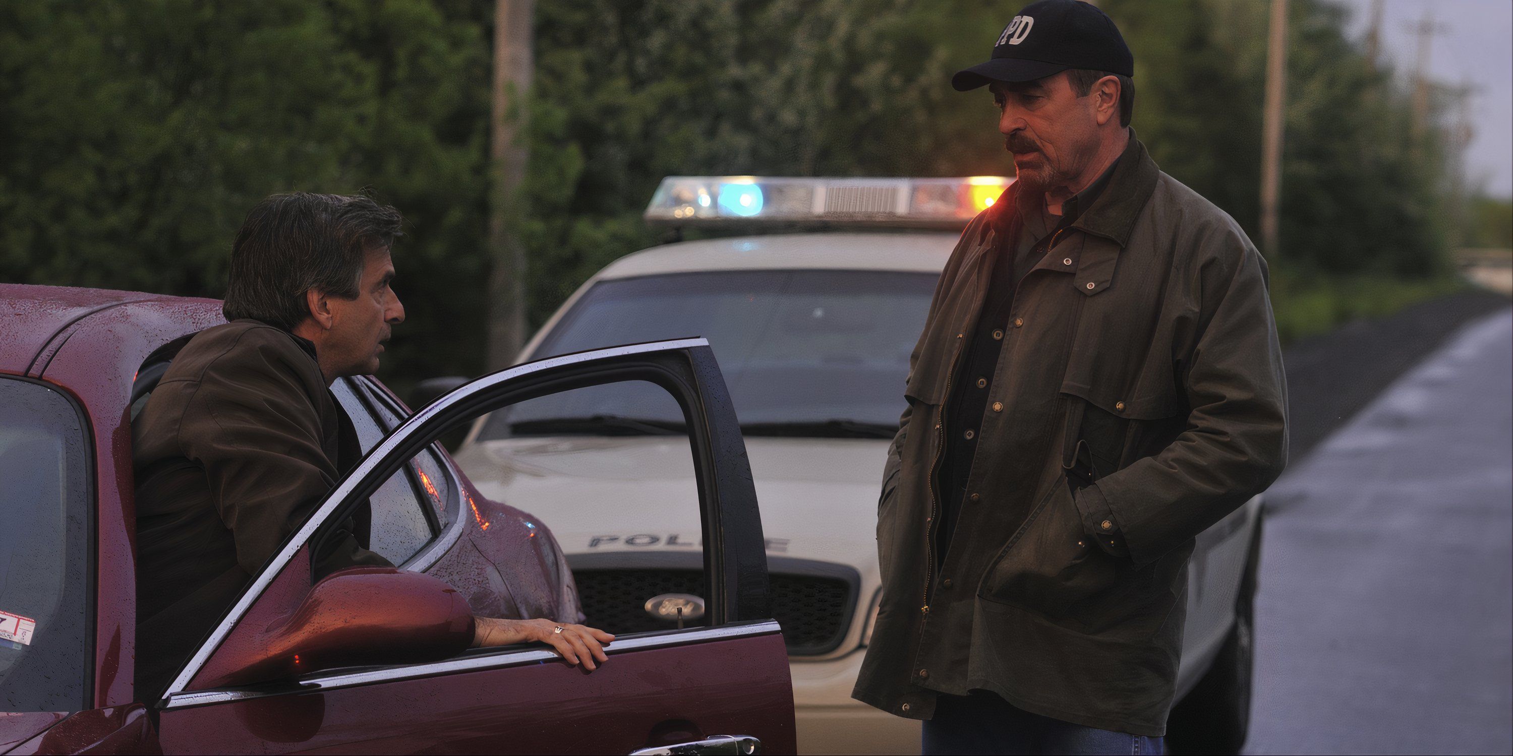 Tom Selleck as Jesse Stone standing on a road talking a man getting out of his car in 'Jesse Stone: Benefit of the Doubt.'