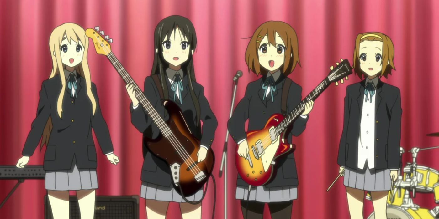 Four female students stand onstage in matching school uniforms holding musical instruments in K-ON! 