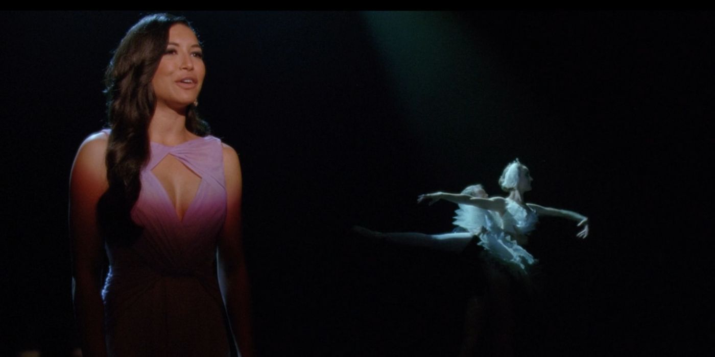 Santana, wearing a purple dress, stands in a spotlight on stage. Behind her, two ballerinas, wearing costumes from Swan Lake, perform a ballet number.
