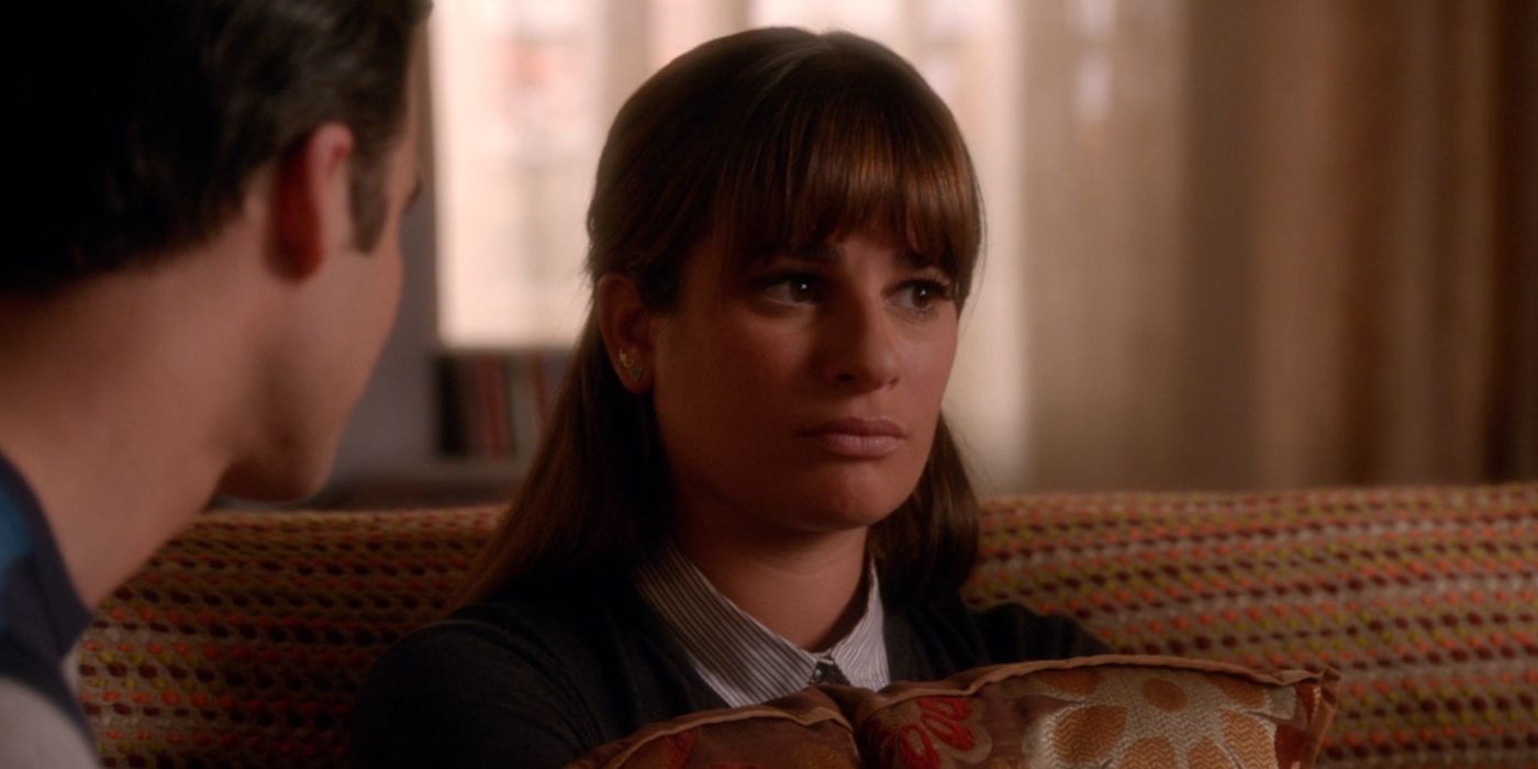 Rachel holds onto a flowery brown pillow. She is sitting with Blaine and her father on a brown sofa covered with a yellow, orange, and green dotted blanket.