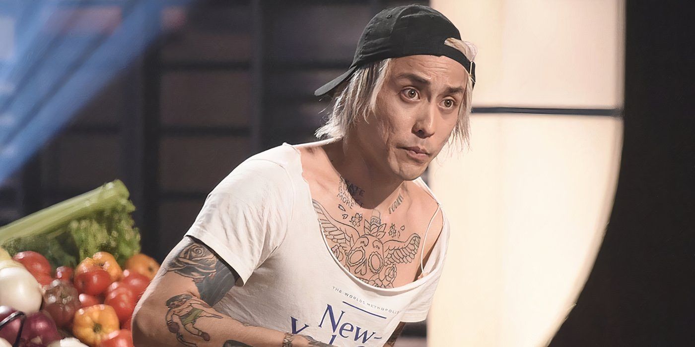Dino from Masterchef leaning forward wearing a scoop neck T shirt showing tattoos on his arms and chest with a backwards hat.