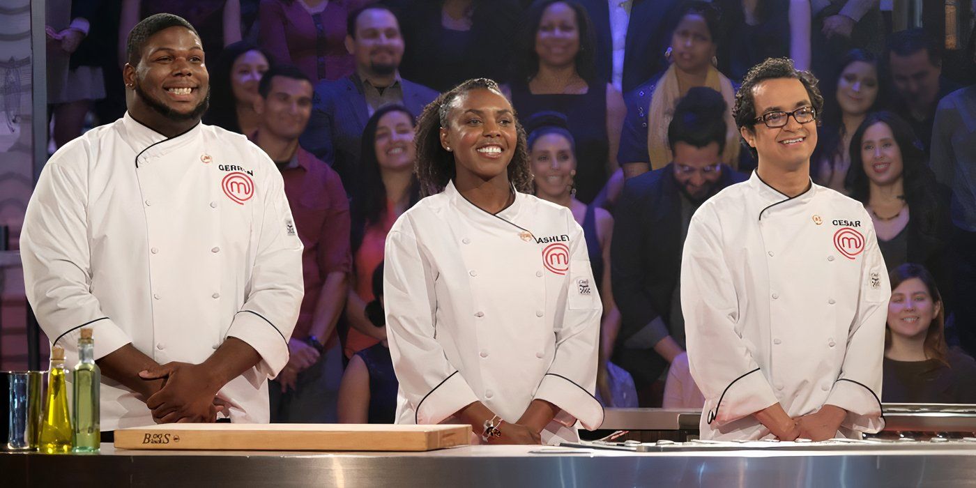 Three contestants smiling wearing white jackets, standing behind a table with an audience behind them on Masterchef.