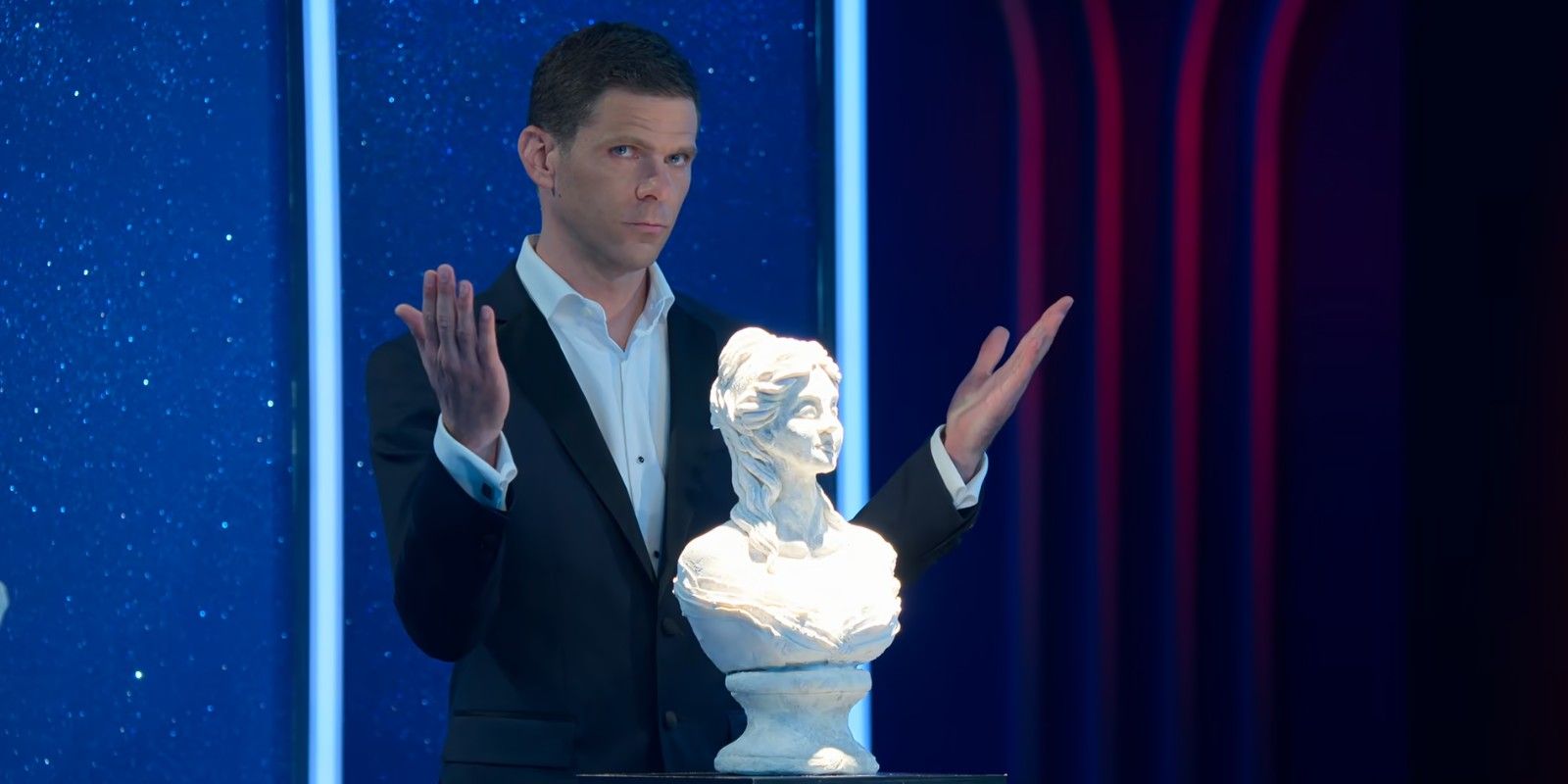 Mikey Day stands behind a lit-up bust of a woman with his hands up - Is It Cake