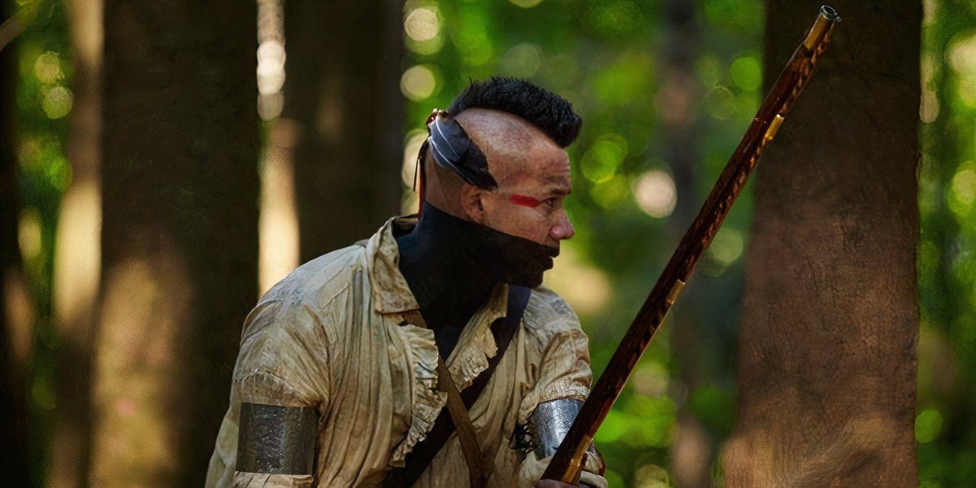 A Mohawk warrior with a painted face and mohawk hairstyle crouches in the woods with a musket