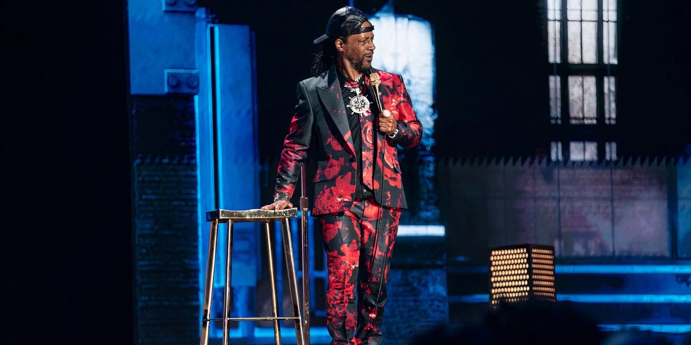 Katt Williams wearing a red and black suit standing on stage, touching a red stool.