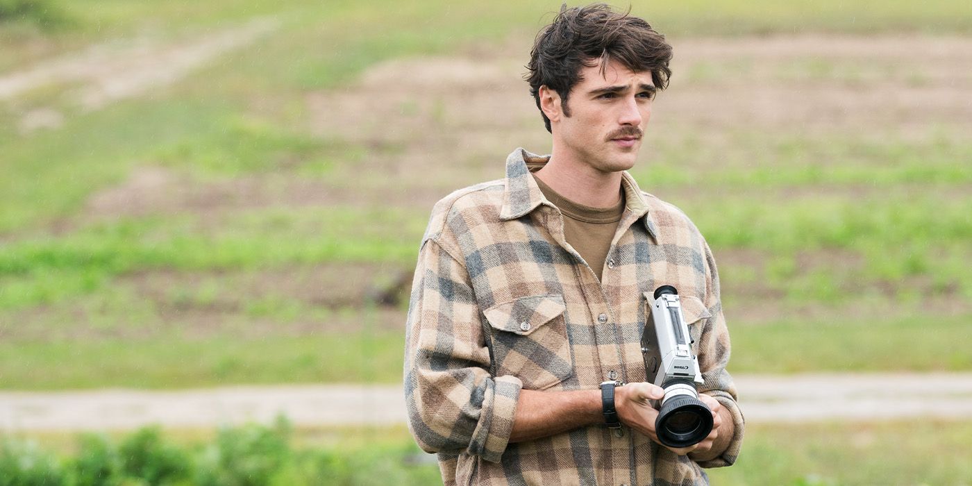 Jacob Elordi with a mustache in plaid on a field holding a camera
