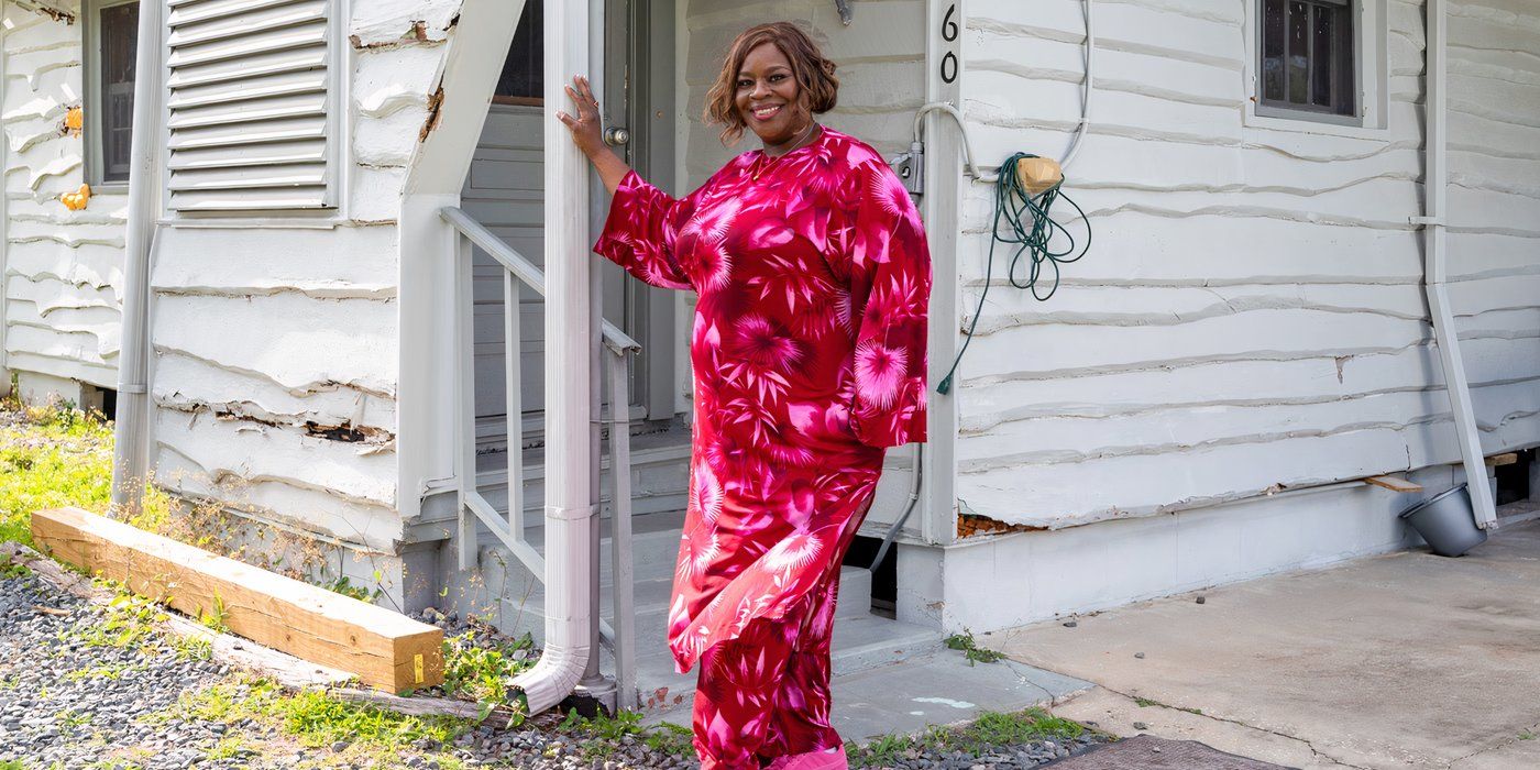 Retta, wearing a pink dress in front of a shabby white house, in Ugliest House in America HGTV